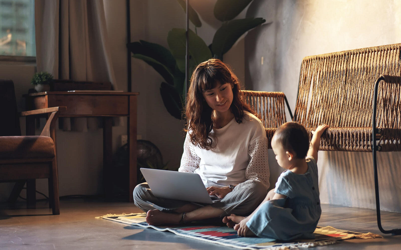 A female professional with a laptop on her lap, sitting on the floor of the lounge and looking at her young child with a big smile on her face.