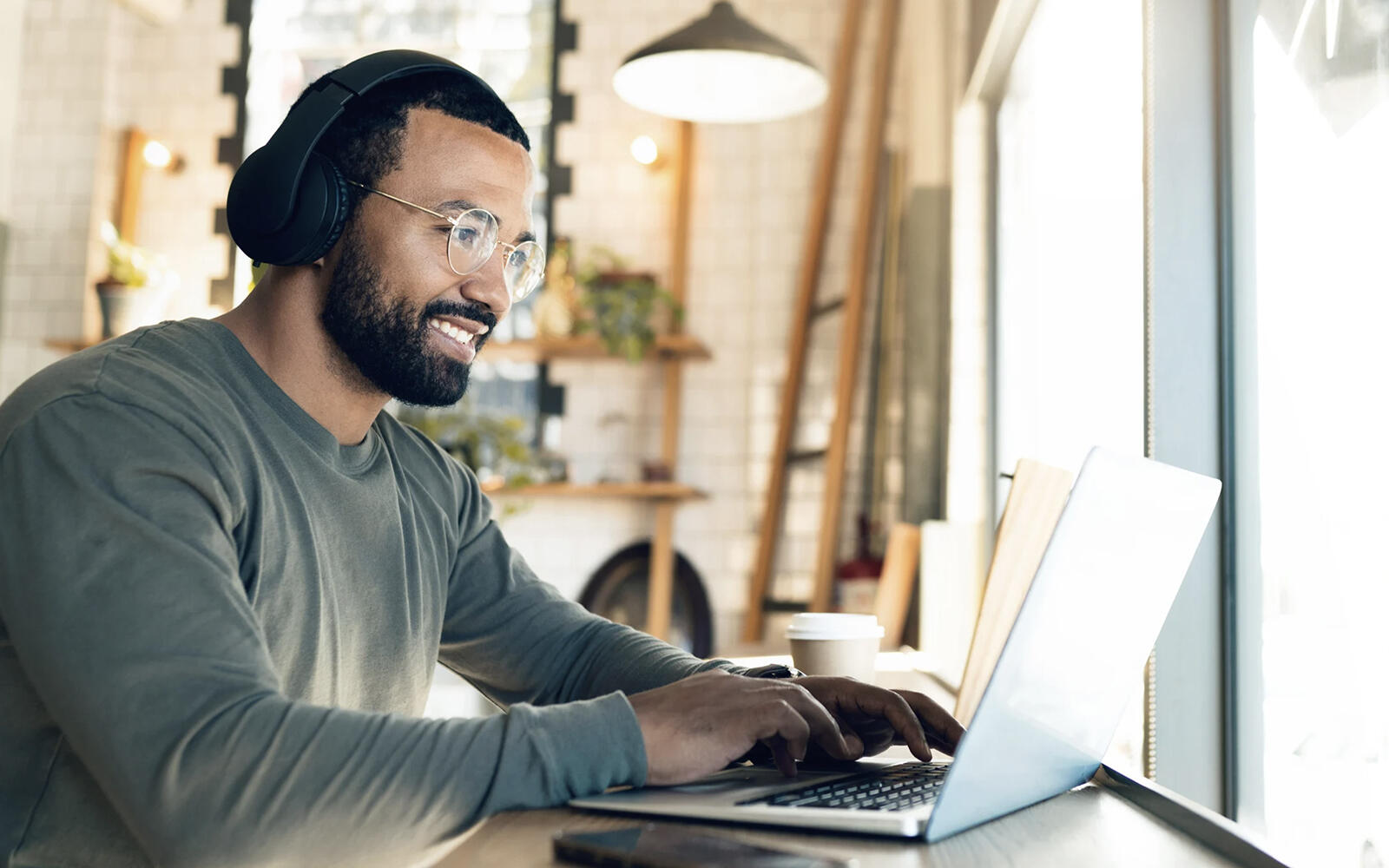 A male professional, wearing glasses and headphones, smiles while working on his laptop in a well-lit office space.