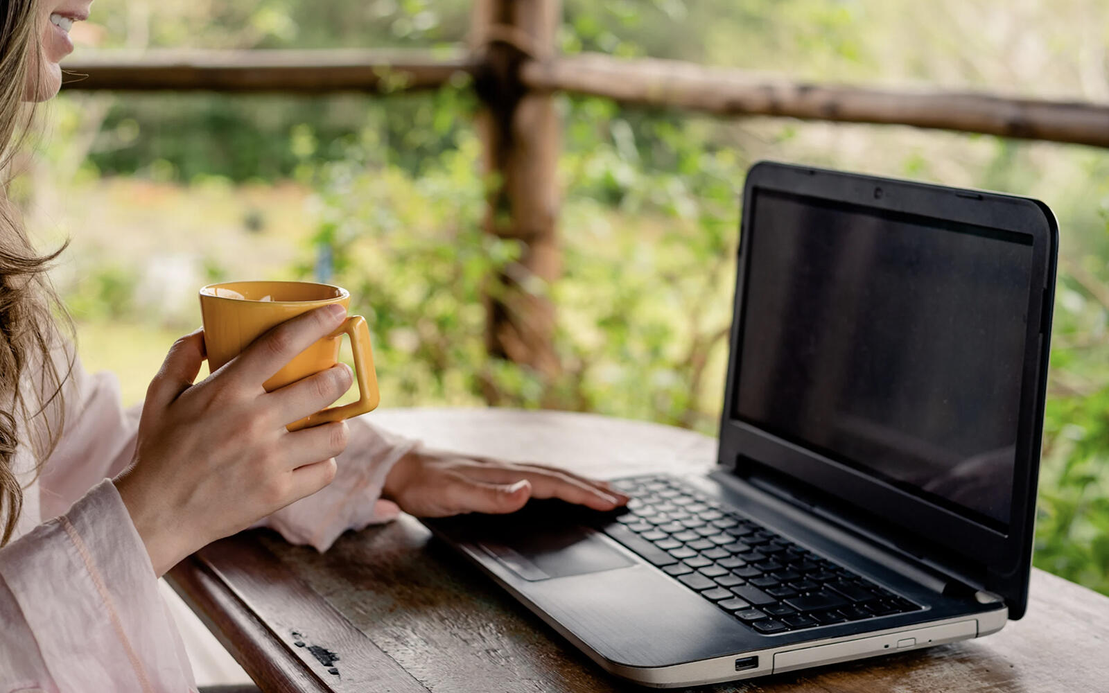 A female professional holding a yellow coffee mug while working on her laptop on the patio of a cabin, surrounded by nature.
