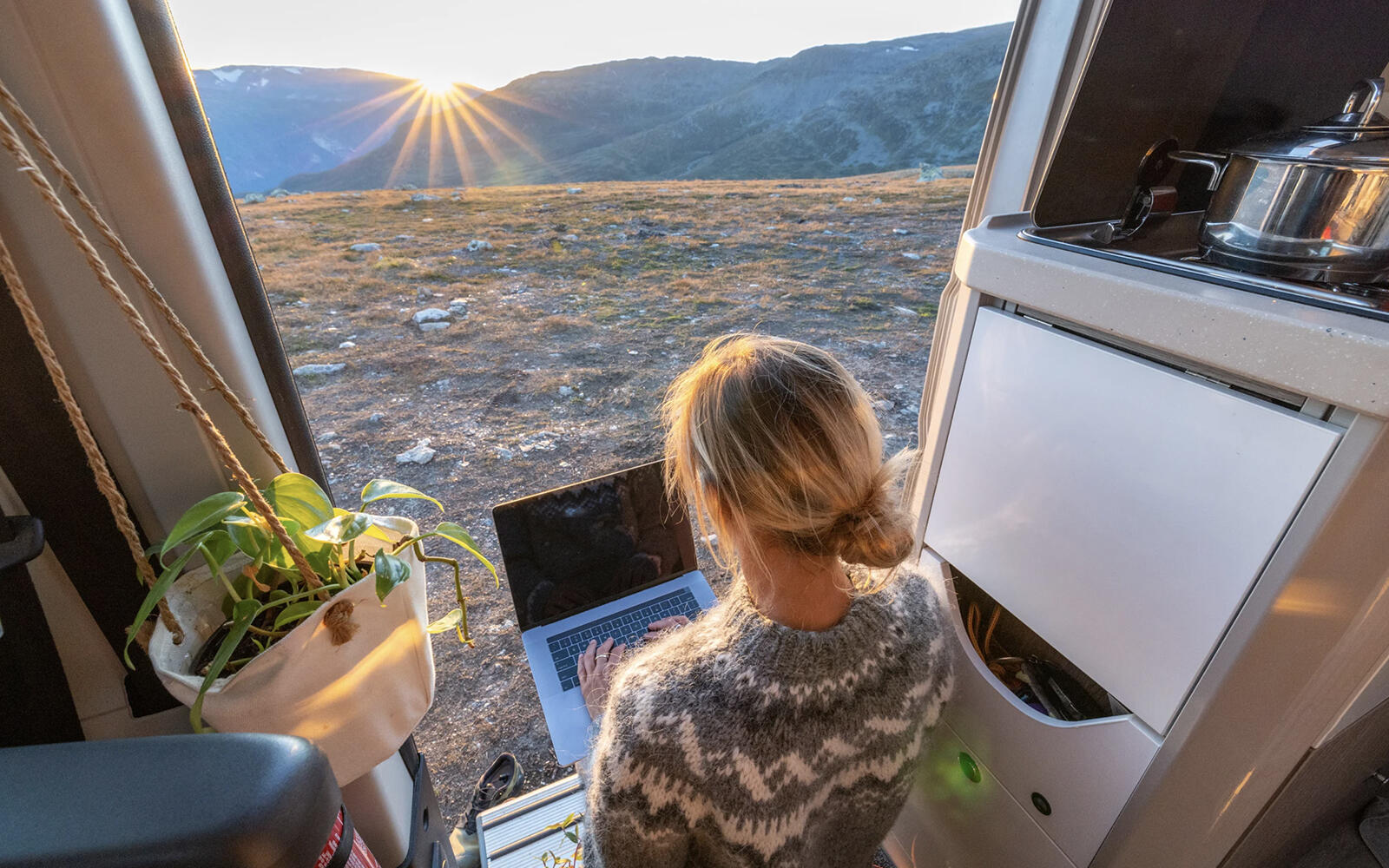 A female professional sitting on the stairs of her caravan, with a laptop on her lap, facing outside towards the mountains during sunset.